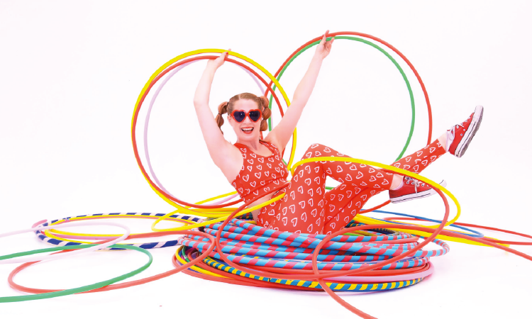 Carla, a white lady with red hair, is lying on a huge stack of hula hoops, in different sizes. She is wearing red sunglasses and holding up different sized hoops