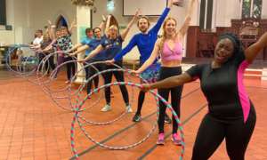 the non-physical benefits of hooping: A group of smiley hoopers line up on the diagonal, with their hoop to one side and their arms in the air!